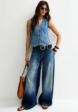 Vrouw die een mouwloze spijkervest en wijd uitlopende verwassen jeans draagt, met een bruine handtas, grote zonnebril en meerdere gestapelde armbanden.