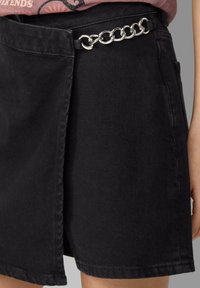 Schwarzer Denim-Rock mit einer seitlichen Tasche und einem metallischen Kettenakzent am Bund. Gerader Schnitt mit fransigem Saum für zusätzliche Textur.