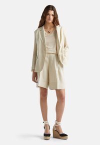 Lnianowy garnitur w jasnym beżu, składający się z marynarki z pojedynczym guzikiem oraz dopasowanych, plisowanych szortów. Połącz go z czarnymi espadrylami i wiązaniami na kostce.