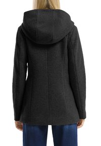 Femme portant une veste à capuche noire texturée et un jean bleu, vue de dos avec les bras détendus le long du corps.