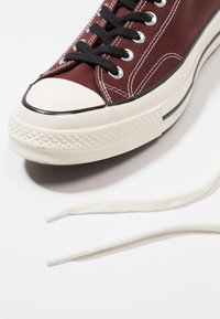 Zapatillas de lona burdeos con cordones negros, suela de goma blanca y capuchón en la punta. Presenta ojetes plateados y costuras blancas contrastantes.