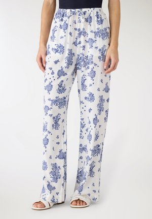 Pantaloni bianchi a gamba larga con motivi floreali blu, vita con coulisse, indossati con sandali bianchi e una maglia nera.