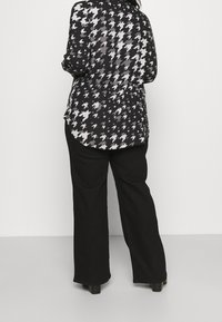 Schwarze und weiße Bluse mit Hahnentrittmuster und abgerundetem Saum, kombiniert mit lockeren schwarzen Hosen und klobigen schwarzen Schuhen.