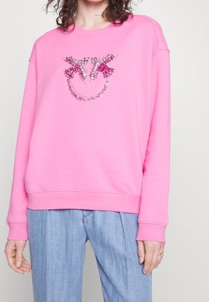 Personne portant un sweat-shirt rose avec deux oiseaux roses formant un motif en forme de sourire en sequins sur la poitrine, associé à un jean bleu clair.