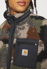 Chaleco de forro polar con patrón de camuflaje en marrón, verde y gris. Cuenta con un bolsillo negro con cremallera y el logotipo de Carhartt, además de un cuello alto.