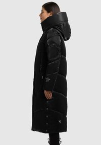 Opgeblazen zwarte donsjas met een hoge kraag en capuchon. Voorzien van een gewatteerd ontwerp, zakken met rits en drukknopen bij de zoom.