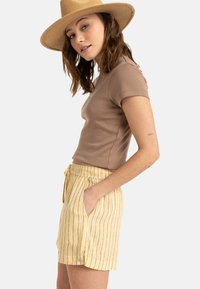 Tricou maro, ajustat, asortat cu pantaloni scurți galbeni cu dungi, care au un tiv elastic și buzunare laterale, model purtând o pălărie cu boruri late.