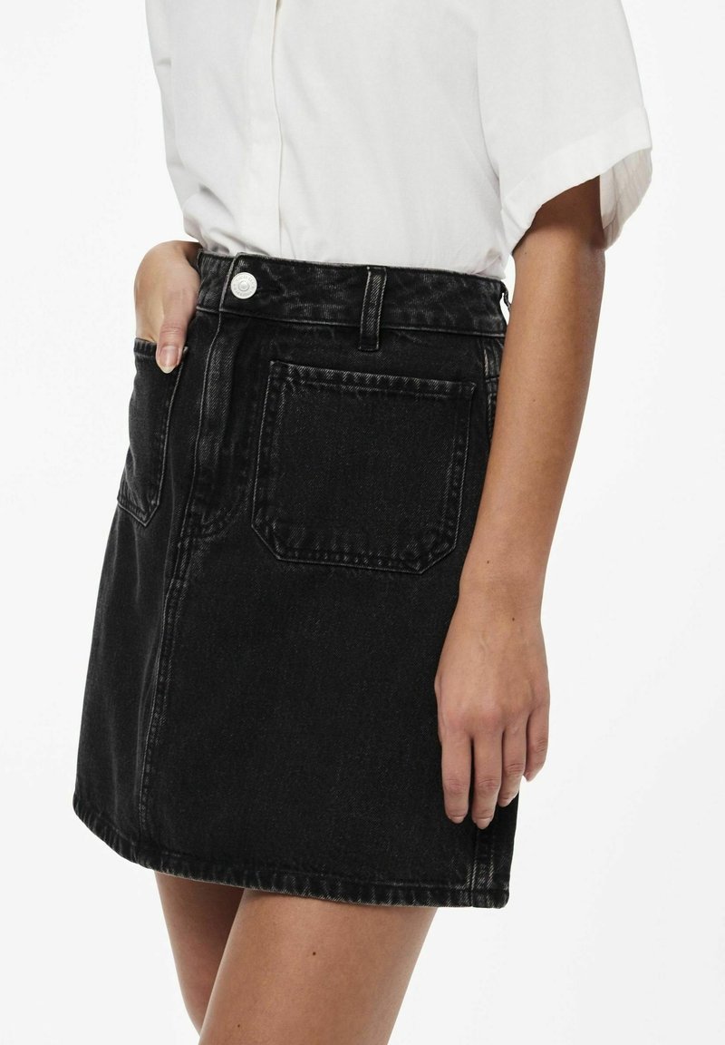 Femme portant une jupe en jean noire avec de grandes poches et une chemise blanche à manches courtes, montrant la main reposant dans la poche et le long de la jupe.