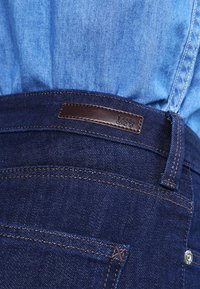 Vaqueros de mezclilla azul oscuro con una etiqueta de cuero marrón que presenta el logo "Lee" en relieve. Costura expuesta y cierre de botón en la cintura.