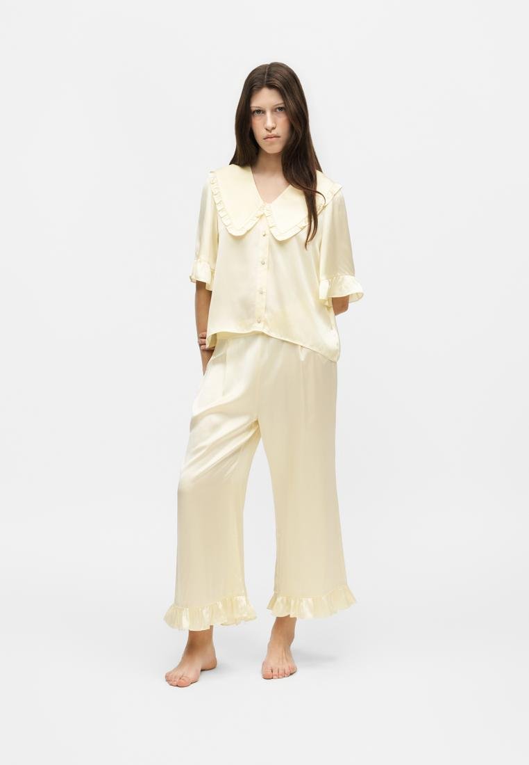 Femme debout, pieds nus, portant un ensemble de pyjama en satin couleur crème avec un col à volants oversize et un pantalon à ourlet volanté, sur un fond uni.