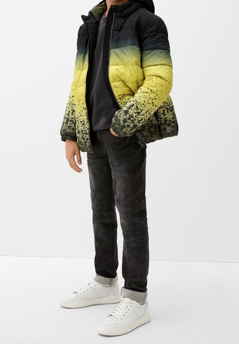 Enfant portant une doudoune noire tachetée de jaune et de vert, un jean foncé aux ourlets retroussés, un t-shirt gris et des baskets blanches, debout les mains dans les poches.