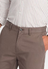 Bruine katoenen broek met een knoopsluiting, zijzakken en een gladde textuur. Draagt een lichtgestreept, collared shirt.