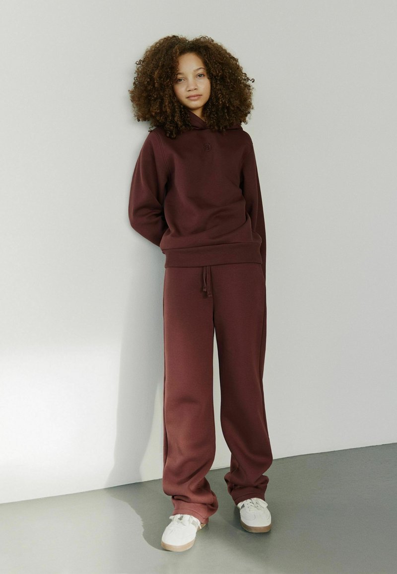 Junge Person mit lockigem Haar, die einen braunen Kapuzenpullover und passende Sweatpants trägt, lässig gegen eine neutrale Wand und einen neutralen Boden stehend.