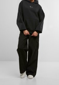 Felpa nera oversize con tasca frontale e logo bianco, abbinata a pantaloni cargo neri larghi con tasche laterali e sneakers bianche.