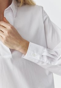 White button-up shirt with a collar and single-button cuffs. Smooth texture, slightly relaxed fit, featuring a placket along the front.