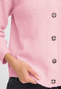 Cardigan tricotat roz cu textură în dungi, cu deschidere pe butoane ce prezintă butoane cu model și mansete ajustate. Material moale și confortabil.