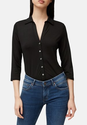 Kobieta ubrana w dopasowaną czarną koszulę na guziki z rękawami do pół łokcia, wpuszczoną w niebieskie dżinsy z średnim stanem, na tle jednolitego tła.