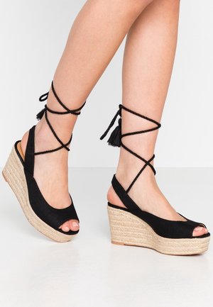 Czarne sandały na koturnie z espadrylową podeszwą, z odkrytymi palcami i krzyżującymi się zapięciami na kostkach ozdobionymi frędzlami. Teksturowana powłoka.