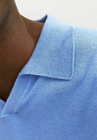Ljusblå polo med en mjuk textur, med en standard krage och en sömlös stickdesign, närbild av halsringningen.