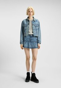 Tânără cu păr scurt blond, îmbrăcată cu o jachetă din denim scurtă, fustă din denim, pulover gri, șosete albe și botine negre cu șireturi.