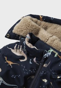 Geacă căptușită dintr-un material bleumarin, cu modele de dinozauri în culori variate. Căptușeală moale din fleece bej și fermoar frontal cu închidere prin capse.