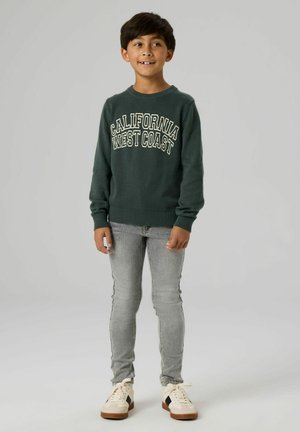 Mørkegrønn genser med teksten "CALIFORNIA WEST COAST" i kontrasterende hvitt, kombinert med grå slanke jeans og hvite joggesko.