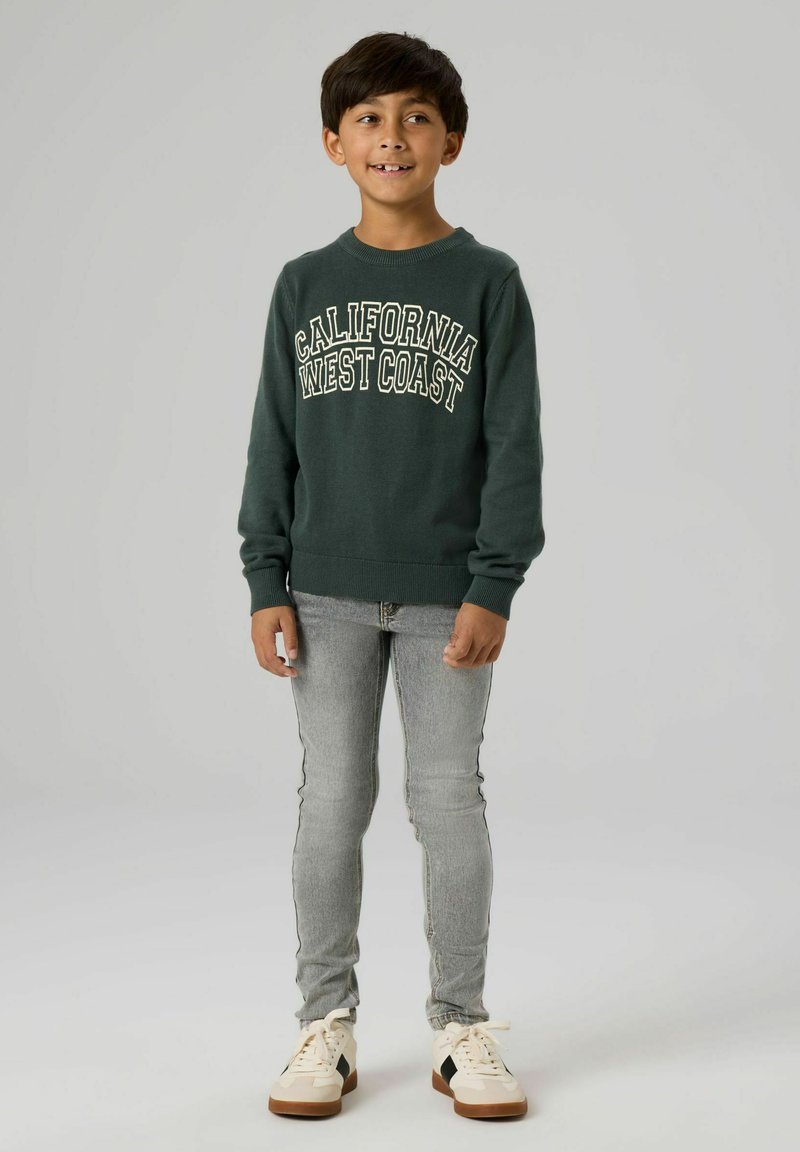 Tamnozeleni duks s natpisom "CALIFORNIA WEST COAST" u kontrastnoj bijeloj boji, uparen sa sivim uskim trapericama i bijelim tenisicama.