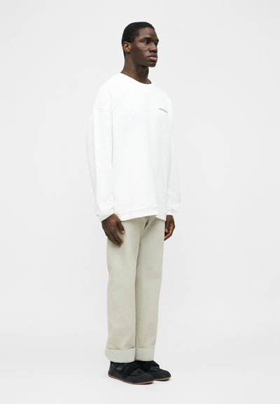 Άνδρας που στέκεται προς τα δεξιά φορώντας λευκό φαρδύ φούτερ, μπεζ παντελόνια με φαρδιά μπατζάκια και μαύρα αθλητικά παπούτσια με λουράκια βέλκρο.