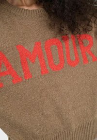 Brūns džemperis ar sarkano tekstu "AMOUR". Tam ir ribota apkakle un apakšmala, izgatavots no teksturētas adījuma materiāla.