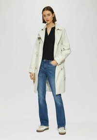 Lichtbeige trenchcoat met dubbele knoopsluiting, gecombineerd met een donkere spijkerbroek en witte sneakers. De jas heeft een kraag en zakken.