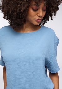 Top corto azzurro chiaro a maniche corte con un motivo ondulato texturizzato. Collo rotondo e vestibilità comoda. Tessuto liscio con una caduta morbida.