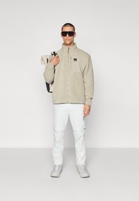 Jachetă din fleece bej cu fermoar pe față și buzunar pe piept, asortată cu pantaloni cargo albi și încălțăminte gri. Modelul ține o geantă.