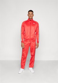 Survêtement en satin rouge avec une veste zippée et un pantalon assorti. Présente des rayures latérales bleues avec des logos et des poignets côtelés. Porté avec des baskets blanches.