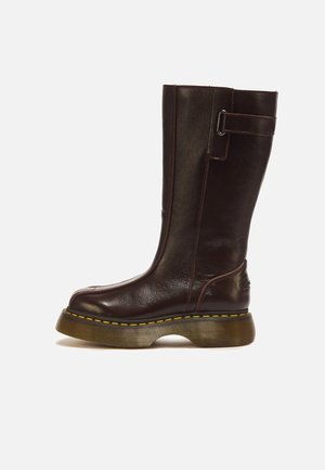 Botte en cuir brun foncé haute avec une semelle épaisse surpiquée en jaune et une sangle avec boucle métallique près du haut.