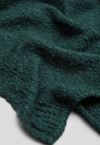 Tissu tricoté vert foncé avec des boucles texturées similaires au bouclé et des plis doux sur un fond clair.