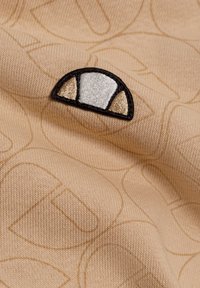 Tela texturizada beige con un patrón sutil, que presenta un logo bordado circular en blanco, negro y acentos en dorado.