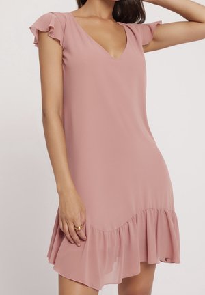 Kobieta nosi krótką, jasnoróżową sukienkę z falbankami na rękawach, dekoltem w kształcie litery V oraz falbanistym dołem, stoi z uniesionym jednym ramieniem.