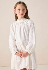 Jeune fille aux cheveux longs portant une robe blanche à manches longues texturée, se tenant avec les mains jointes devant un fond uni.