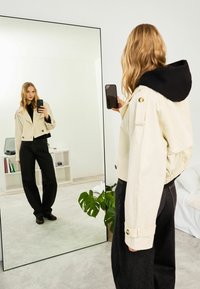 Veste courte couleur crème avec un sweat à capuche noir en dessous, associée à un pantalon noir ample. Arrière-plan miroir réfléchissant et plante visible.
