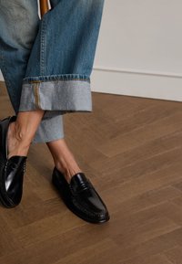 Schwarze Lederl davant mit Stickdetails, kombiniert mit verkürzten blauen Jeans mit umgeschlagenem Saum und Kontrastnähten. Brauner Holzboden.