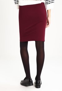 Vinröd figursydd kjol i stretchmaterial med en mjuk yta. Bärs med svarta täckande strumpbyxor och svarta skor, sedd bakifrån.