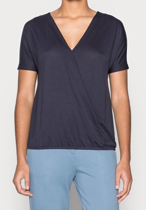 Osoba ubrana w granatowy krótki rękaw, bluzkę z dekoltem w serek w stylu kopertowym oraz jasnoniebieskie spodnie, stojąca na jednolitym tle.