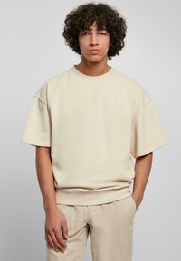 Beige, túlságosan bő pulóver, rövid ujjak, kerek nyakkivágás és ribbelt hem, puha anyagból készült. Hozzá illő bézs nadrággal párosítva.