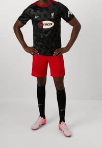 Musta jalkapallopaita punaisilla yksityiskohdilla ja abstraktilla kuviolla, yhdistettynä punaisiin shortseihin ja mustiin sukkiin. Vaaleanpunaiset jalkapallokengät täydentävät asun.