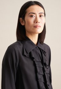 Blusa nera con dettaglio anteriore a volant, colletto strutturato, texture liscia e chiusura a bottoni, presentata su uno sfondo neutro.
