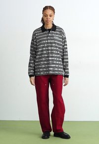 Polo a maniche lunghe a righe bianche e nere con colletto nero, abbinato a pantaloni rossi larghi e scarpe nere. Sfondo semplice.