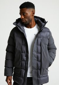 Uomo che indossa un piumino grigio scuro sopra una maglietta bianca con stampa, che guarda a sinistra con le mani nelle tasche del giubbotto.