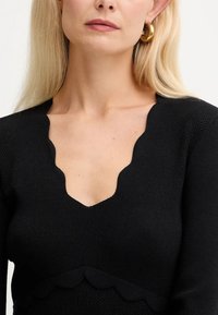 Donna bionda che indossa un top nero a maniche lunghe con scollatura a V ondulata e grandi orecchini a cerchio dorati.