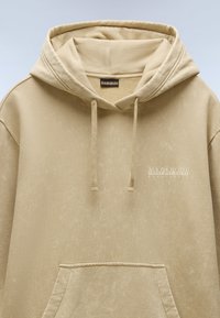 Sudadera beige con acabado texturizado, cordones y bolsillo canguro. Presenta un logotipo impreso en blanco en el pecho. Tejido suave.