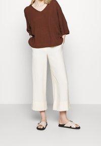 Maglione marrone oversize con scollo a V, abbinato a pantaloni larghi color crema e sandali con incrocio rosa chiaro e suola nera.
